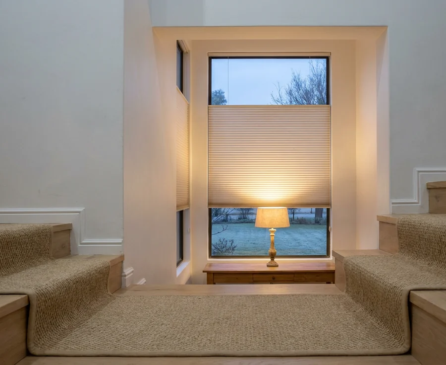 Cellular honeycomb blind lowered two-thirds down the tall stairwell window of a Cornwall Hill home at winter dusk, a brass table lamp on the sill and bare trees on the far side of the glass